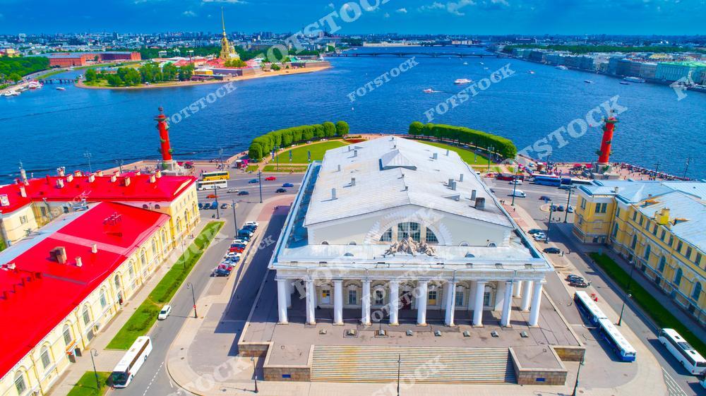 На фотографии изображена Стрелка Васильевского острова с ее знаковыми достопримечательностями — зданием Биржи и Ростральными колоннами. Это сердце исторического Санкт-Петербурга, где сливаются архитектура и водные просторы Невы. На заднем плане возвышается Петропавловская крепость, добавляя акцент петербургскому пейзажу. Яркий солнечный день подчеркивает контраст красных крыш, колонн и голубого неба, придавая этому виду особенную живописность.